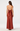 A woman viewed from the back wearing a floor-length rust-brown halter dress with an open back and large bow detail at the waist, photographed against a light gray studio background.